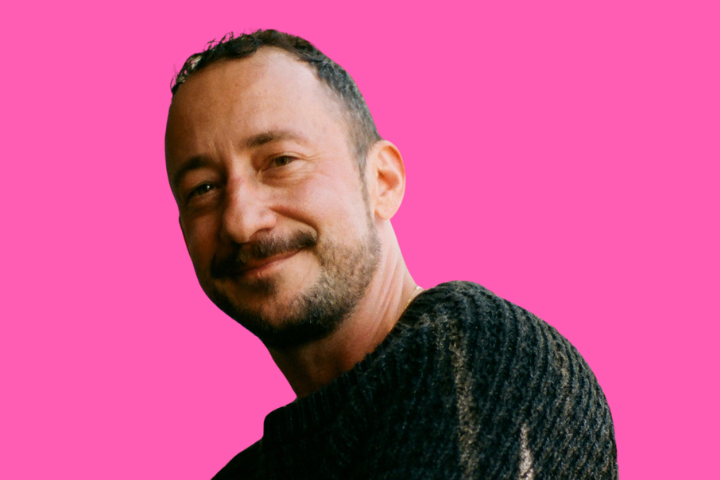 A person with short hair and a beard is smiling while wearing a dark, textured sweater. The background is solid bright pink.