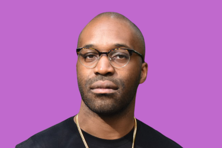 A man with short hair and glasses, wearing a black shirt and gold necklace, stands against a solid purple background.
