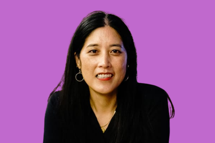 A woman with long black hair, wearing hoop earrings and a black top, smiles in front of a plain purple background.