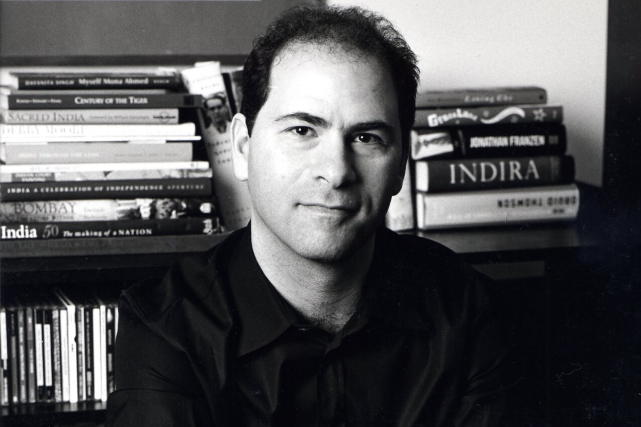 A man with short dark hair wearing a dark shirt sits in front of a bookshelf filled with books and CDs. The image is in black and white.