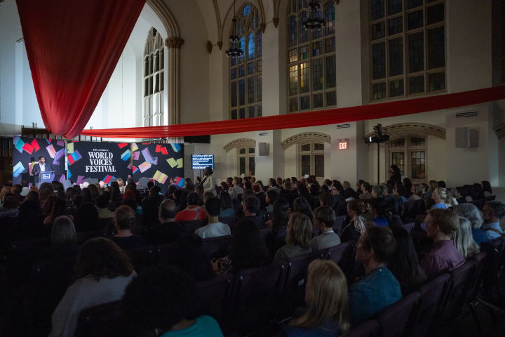 A large audience sits in a dimly lit hall with arched windows, watching a speaker at a podium during the World Voices book literary festival. Colorful banners and red fabric decorate the venue.