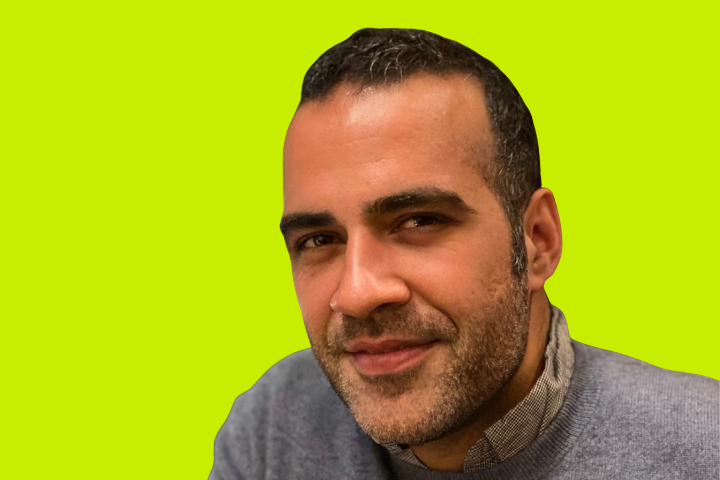 A man with short dark hair and a trimmed beard wears a gray sweater and light shirt, smiling slightly against a bright yellow-green background.