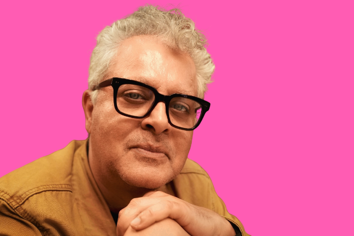 A middle-aged man with gray hair, black glasses, and a tan jacket smiles gently, resting his hands under his chin, against a bright pink background.