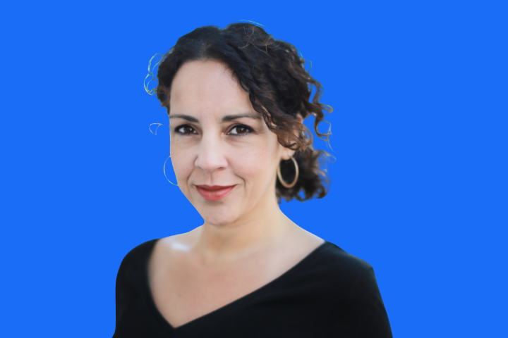 A woman with curly dark hair, wearing large hoop earrings and a black top, stands against a solid bright blue background, smiling slightly at the camera.
