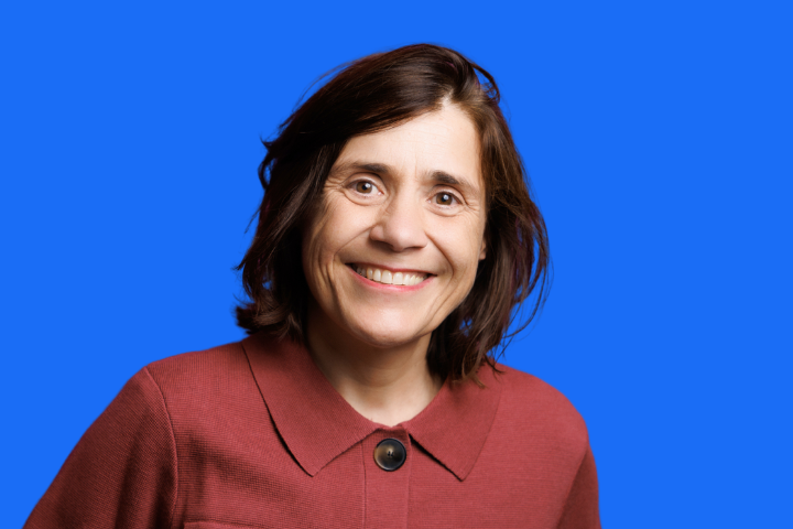 A woman with shoulder-length brown hair, wearing a maroon collared jacket, smiles at the camera against a solid blue background.