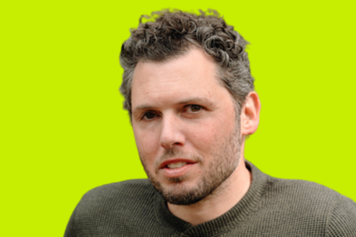 A man with short, curly dark hair and a beard wearing a textured, olive-green sweater, set against a bright yellow-green background.