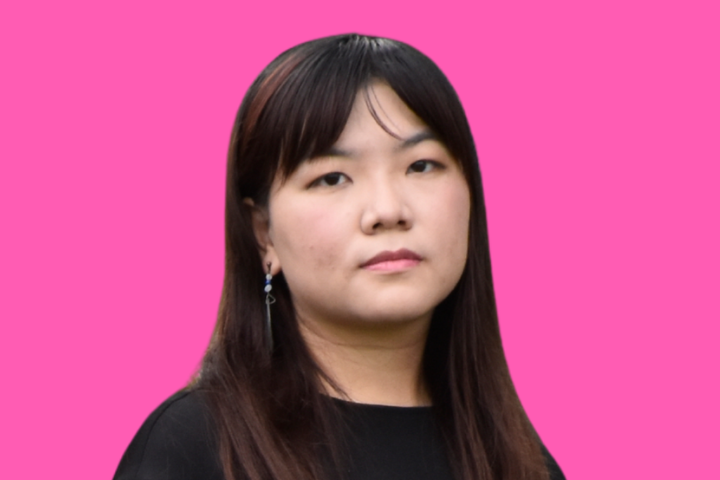 A woman with straight dark hair wearing a black top stands in front of a bright pink background, looking at the camera with a neutral expression.