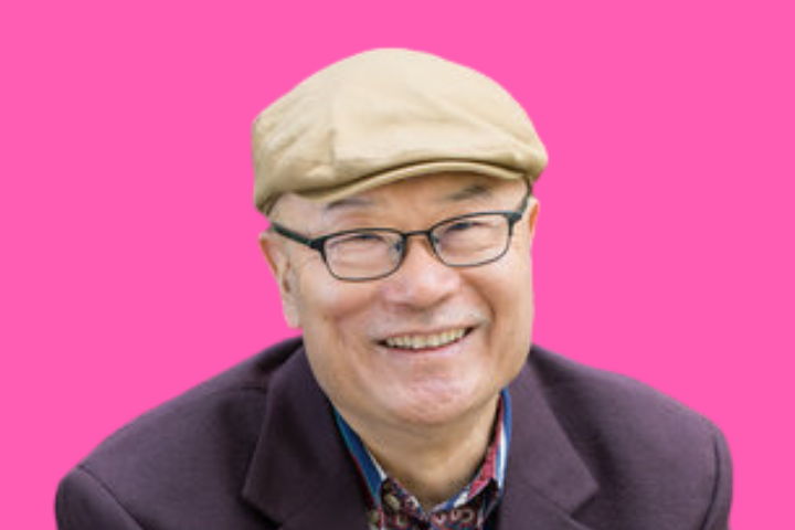 An older man wearing glasses, a tan flat cap, and a dark jacket smiles at the camera in front of a bright pink background.