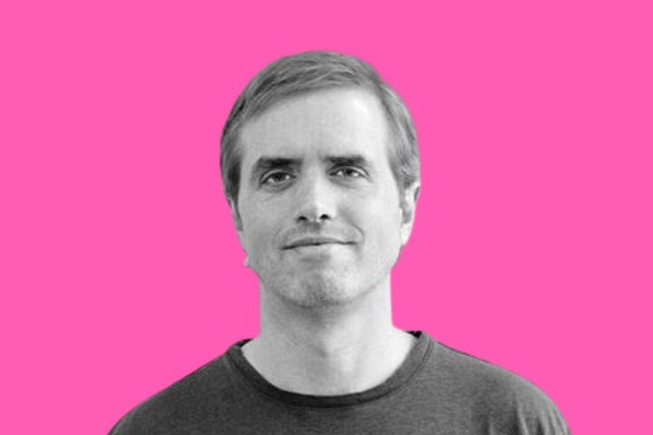 A man with short light hair and a slight smile, wearing a plain dark shirt, is shown against a bright pink background.