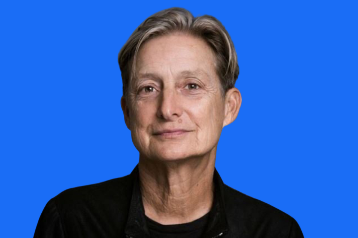 A person with short, light brown hair and fair skin, wearing a black jacket and dark shirt, smiles slightly in front of a solid bright blue background.