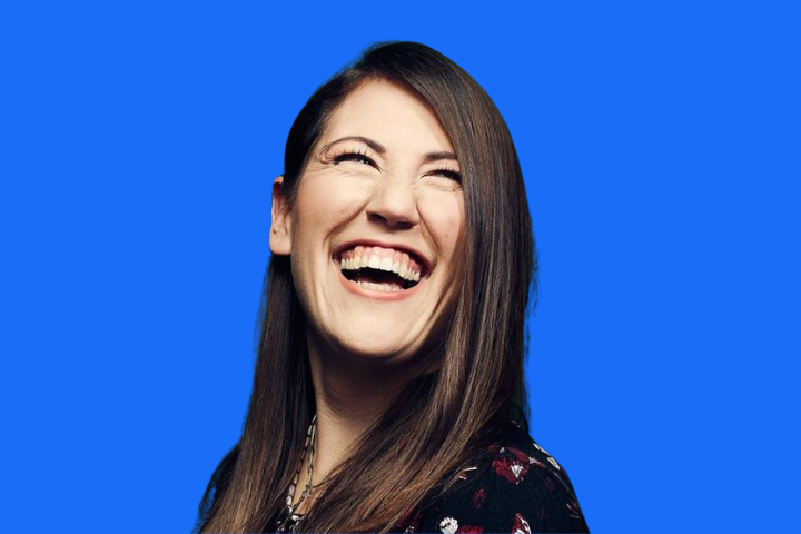 A woman with long brown hair is smiling broadly and looking slightly upward. She is wearing a patterned dark top and standing in front of a solid bright blue background.