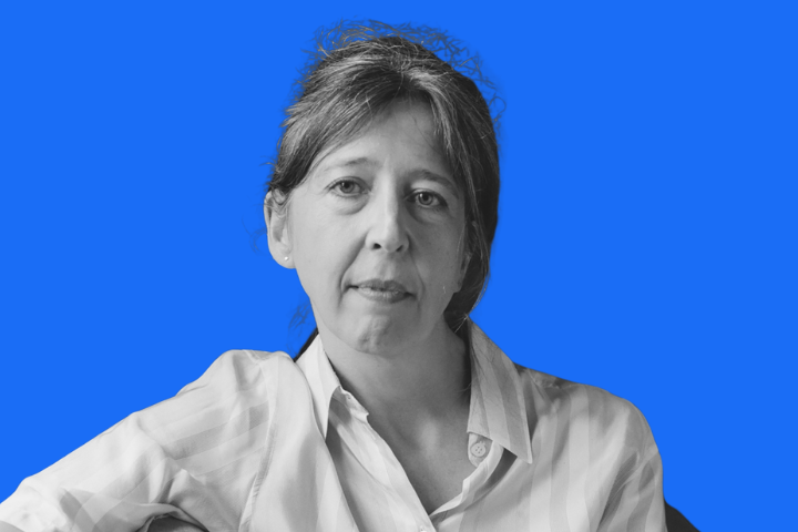 A woman with light skin and brown hair, wearing a light-colored button-up shirt, looks at the camera with a neutral expression. The background is a solid bright blue.