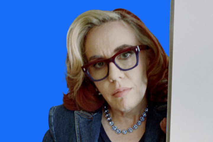 A woman with wavy red and blonde hair, wearing large dark-framed glasses, a denim jacket, and a blue beaded necklace, looks directly at the camera against a bright blue background.