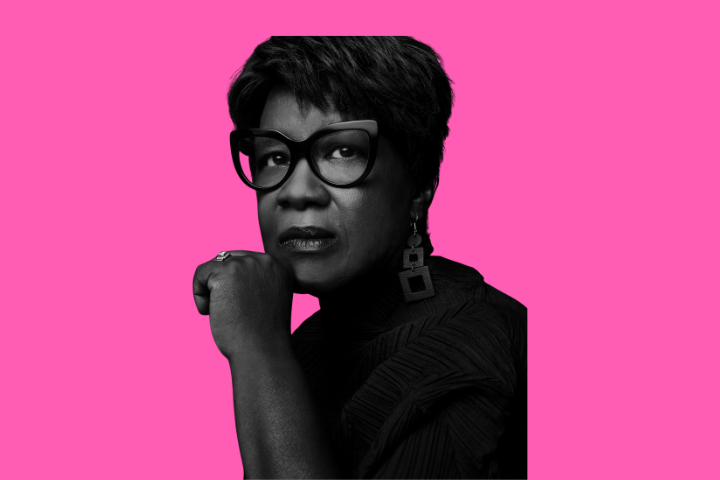 A black-and-white portrait of a woman with short hair, large dark cat-eye glasses, and geometric earrings, resting her chin on her hand, set against a bright pink background.