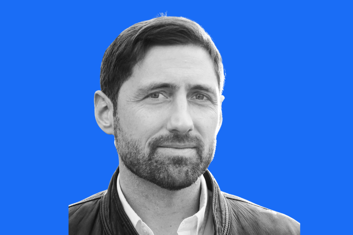 Black and white photo of a man with short dark hair, a beard, and a slight smile, wearing a jacket over a collared shirt, set against a solid bright blue background.