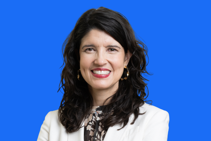 A woman with long dark hair smiles at the camera. She is wearing a white blazer over a floral blouse and gold hoop earrings, standing against a solid bright blue background.