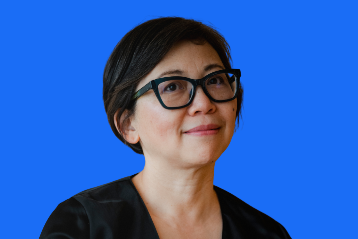 A woman with short dark hair and black glasses, wearing a black top, smiles slightly while looking upward, posed against a solid bright blue background.