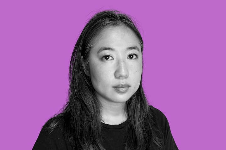 A woman with long dark hair and a neutral expression, wearing a black shirt, is shown against a solid purple background. The photo is black and white except for the background.