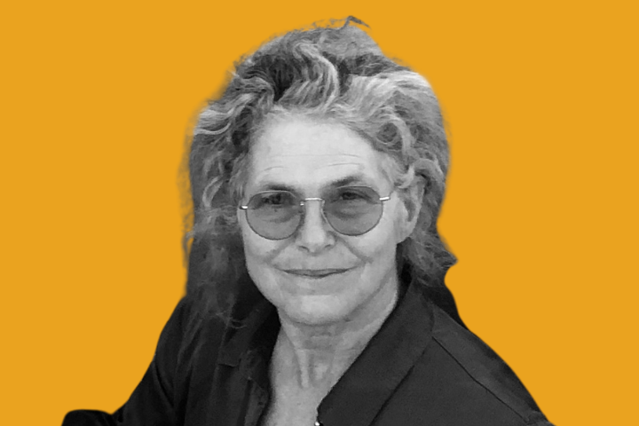 A grayscale portrait of an older person with wavy hair, wearing glasses and a collared shirt, smiles softly against a solid orange background.