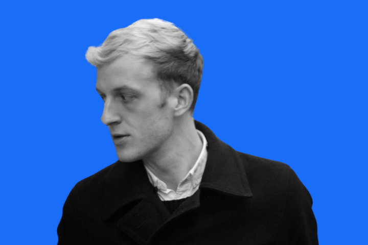 A young man with light hair wearing a dark coat and white shirt looks to his left. The image is in black and white except for a solid bright blue background.