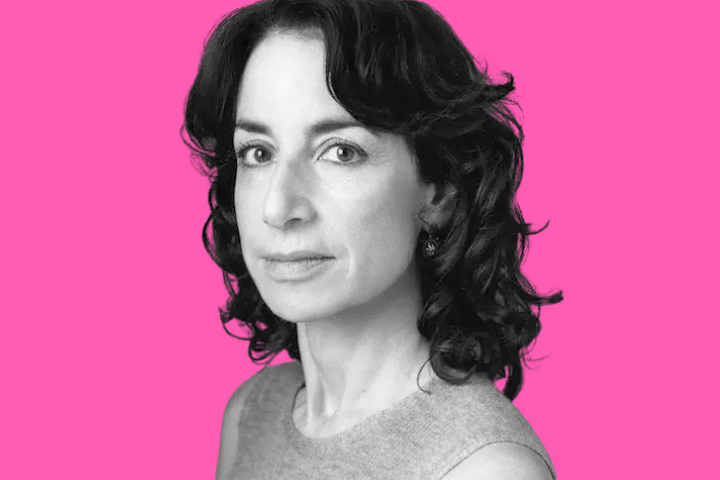 A woman with wavy dark hair and a sleeveless top looks at the camera against a bright pink background. The image is in black and white except for the background.