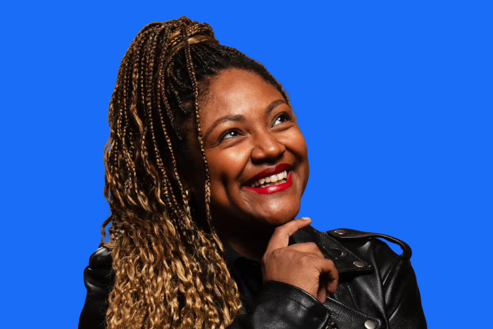 A smiling woman with long, braided hair wearing a black leather jacket poses with her hand on her chin against a solid bright blue background.