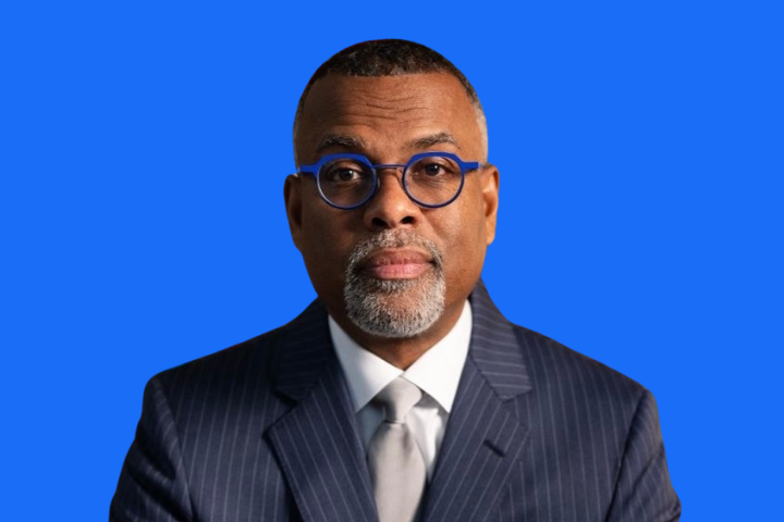 A man in a pinstripe suit, white shirt, and tie, wearing blue-rimmed glasses, poses against a solid blue background.
