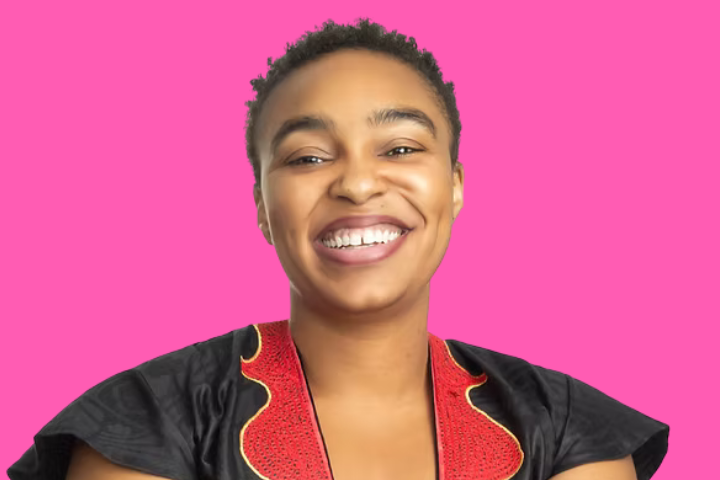 A person with short hair smiles warmly, wearing a black top with a red and gold patterned collar, against a bright pink background.