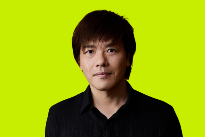 A man with straight dark hair wearing a black collared shirt stands against a bright yellow-green background, looking directly at the camera with a neutral expression.