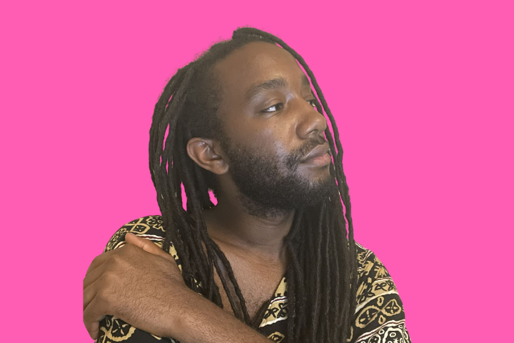 A person with long dreadlocks and a beard looks to the side, with one arm resting on their opposite shoulder, against a bright pink background. They wear a patterned shirt and have a calm, thoughtful expression.