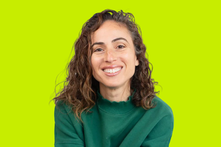 A woman with wavy brown hair wearing a green sweater is smiling at the camera. The background is a bright lime green color.