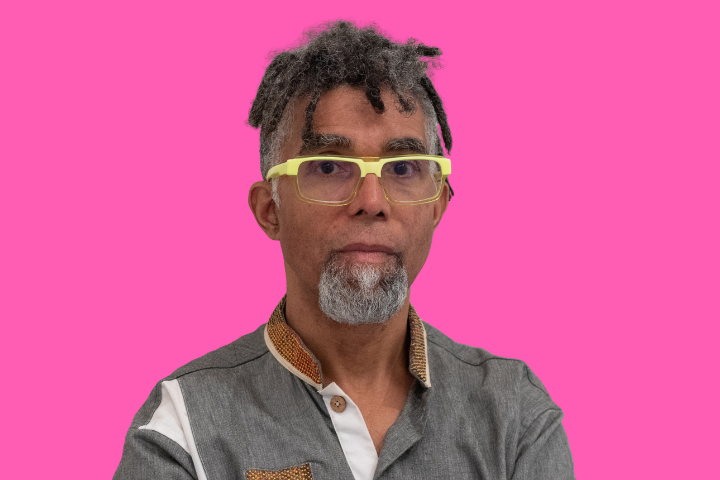 A person with short dreadlocks, gray beard, and mustache, wearing yellow glasses and a gray collared shirt, stands in front of a bright pink background.