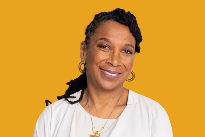 A woman with dark, curly hair styled in twists, wearing hoop earrings and a white top, smiles softly against a solid mustard-yellow background.