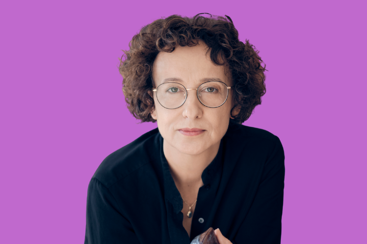 A person with short curly brown hair, wearing round glasses and a dark shirt, looks directly at the camera against a solid purple background.