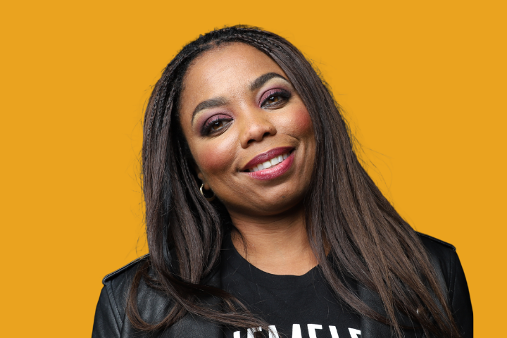 A woman with long brown hair and braids smiles at the camera. She is wearing a black top and a leather jacket, with a golden yellow background behind her.