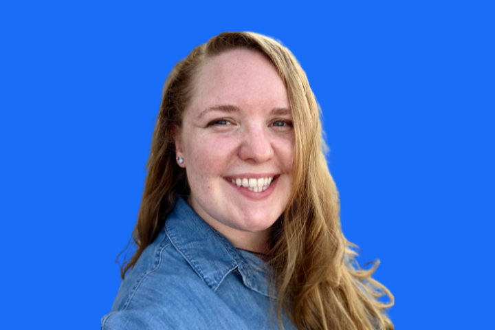 A woman with long, light brown hair smiles at the camera, wearing a denim shirt against a solid bright blue background.