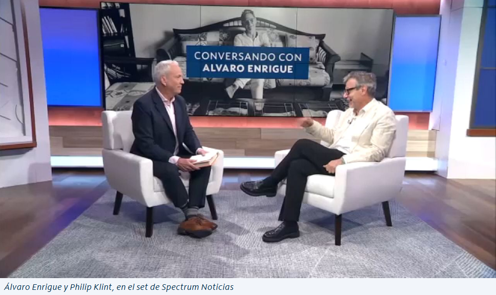 Two men sit facing each other in white armchairs on a TV studio set, discussing press releases and festival highlights. A screen behind them displays “Conversando con Alvaro Enrigue.” Both are engaged in conversation, one holding notes.